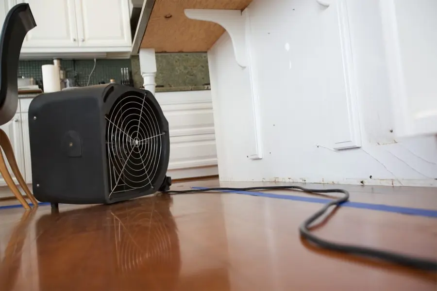 A black air mover on a wood floor beside a kitchen counter, blowing across the boards