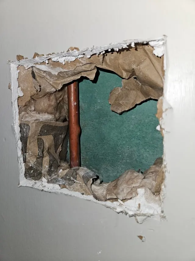 A square hole cut in drywall that exposes a bare pipe inside the wall