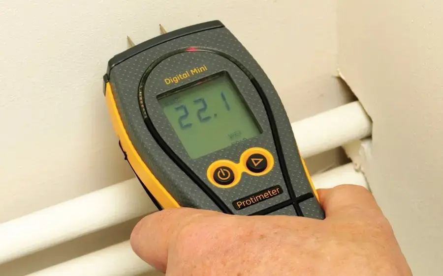 A pin moisture meter held against a Covina wall above two pipes to check if the drywall is still wet.